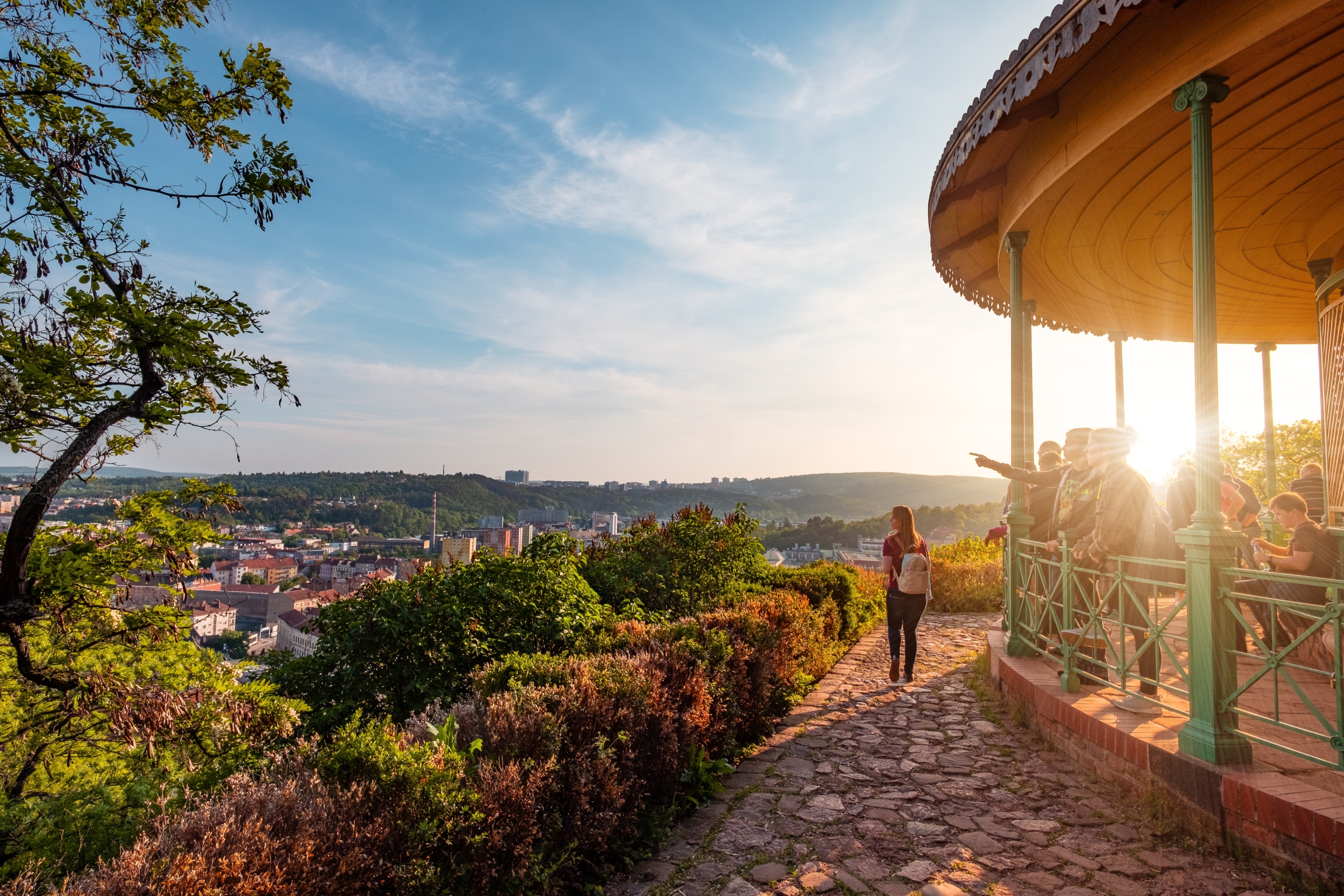 People stand on a stone walkway beside a green ornate gazebo, overlooking a city below under a bright blue sky with sunset light.