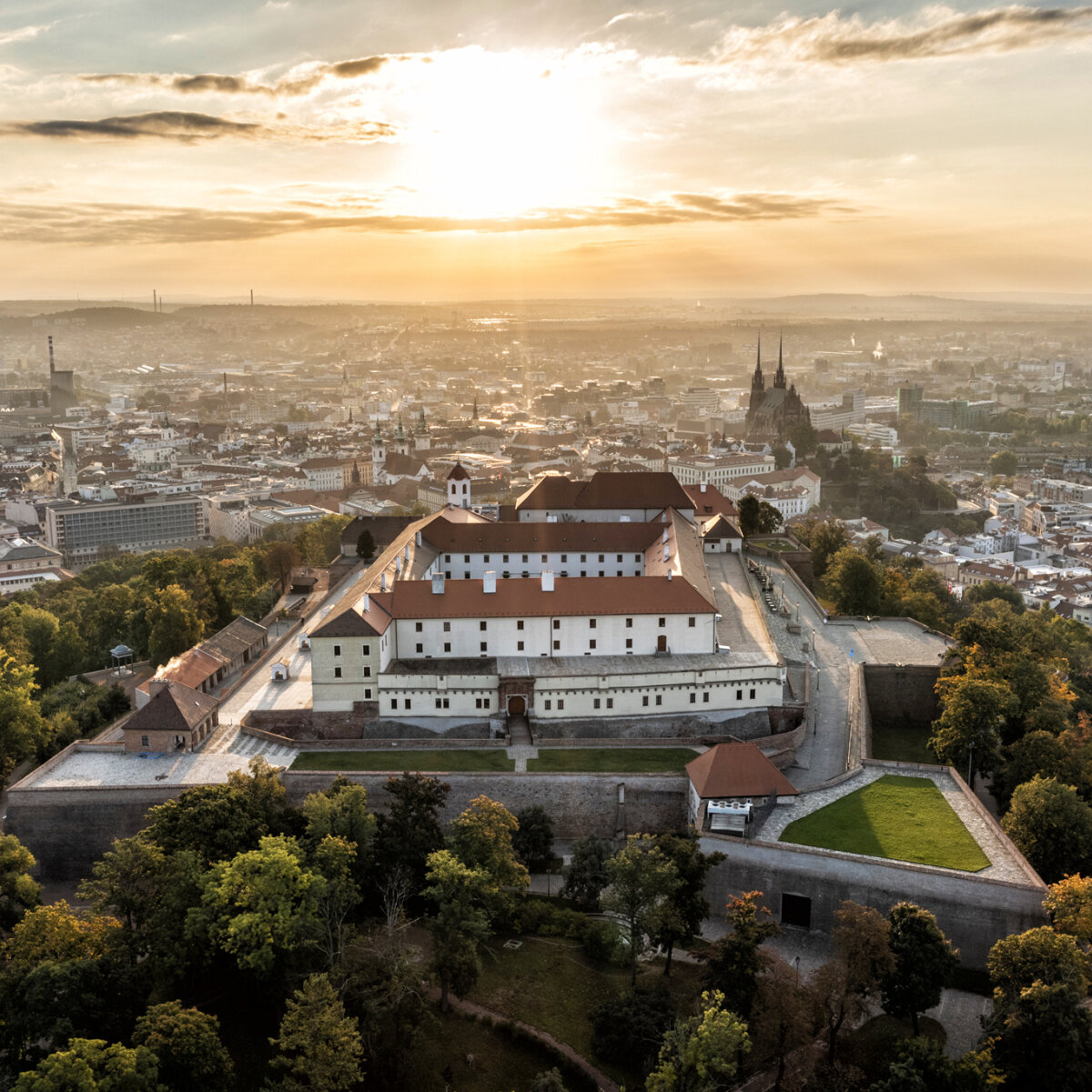 Aerial view of a large hilltop castle with white walls and red roofs overlooking a European city at sunset.