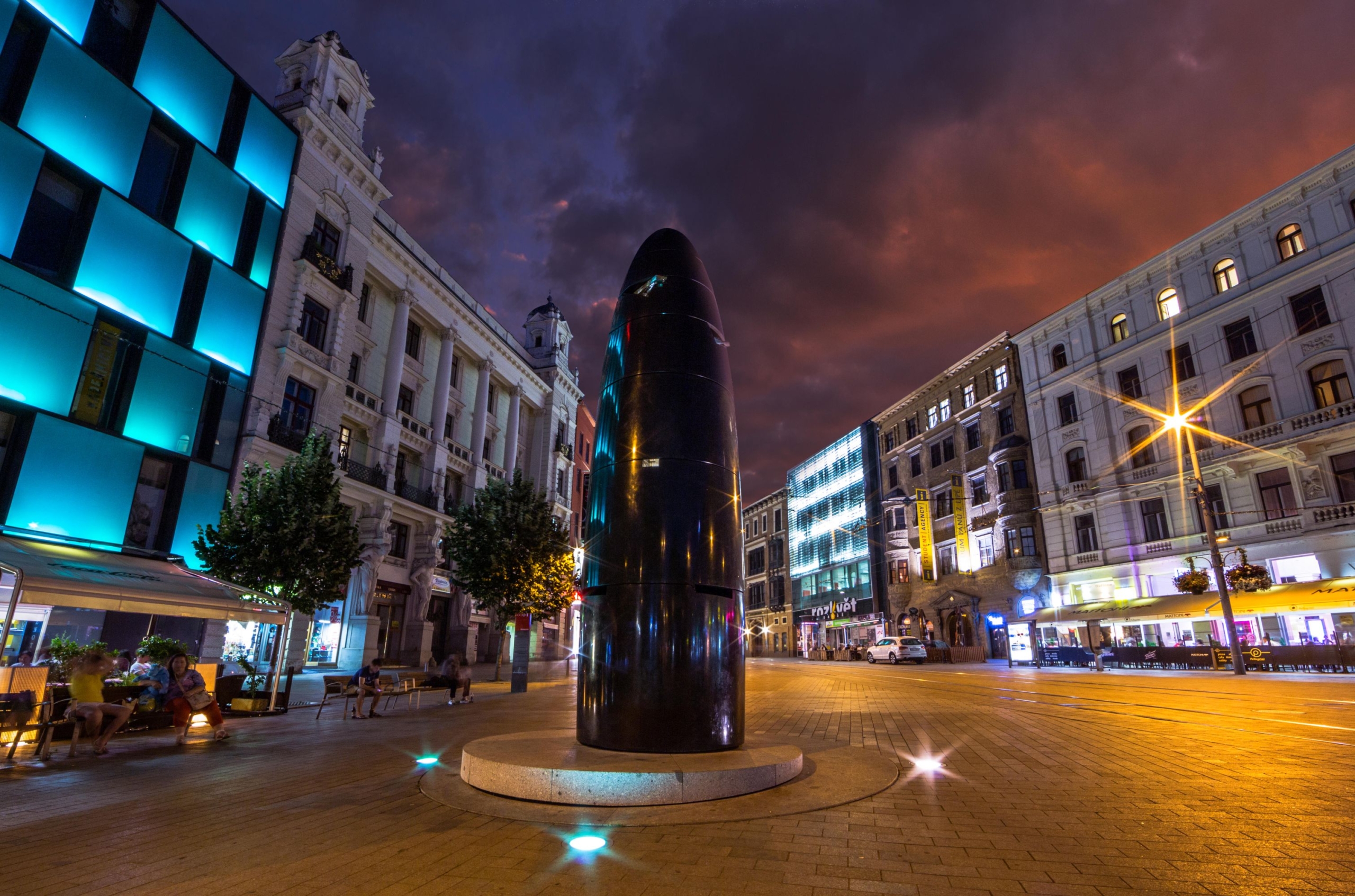 Night city square with a tall glossy black cylindrical monument centered, surrounded by illuminated buildings and a purple-orange sky behind. People sit at outdoor cafes on the left as blue-lit façades glow.