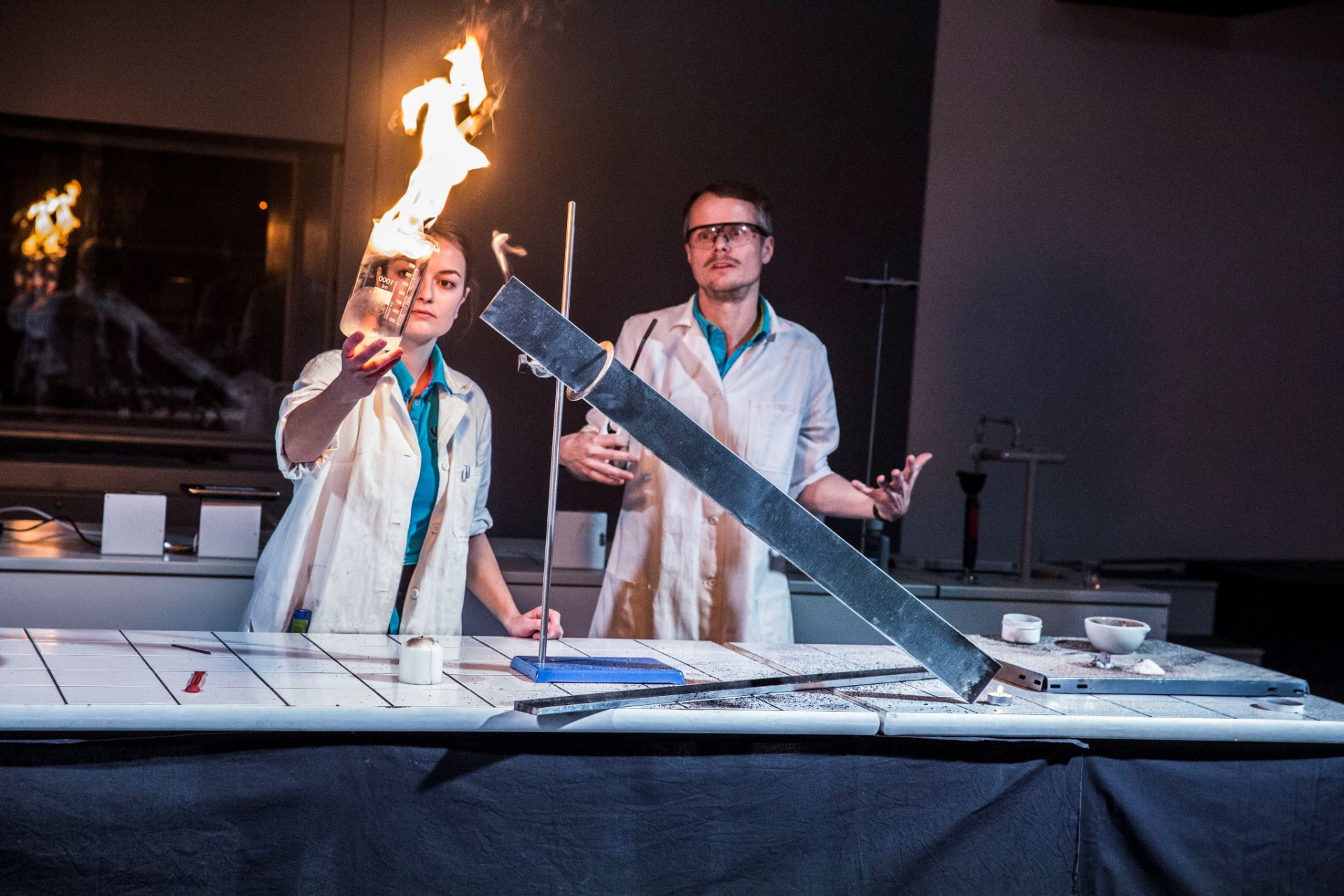 Two scientists in lab coats perform a flame experiment at a lab bench, one holding a burning flask toward the flame source.