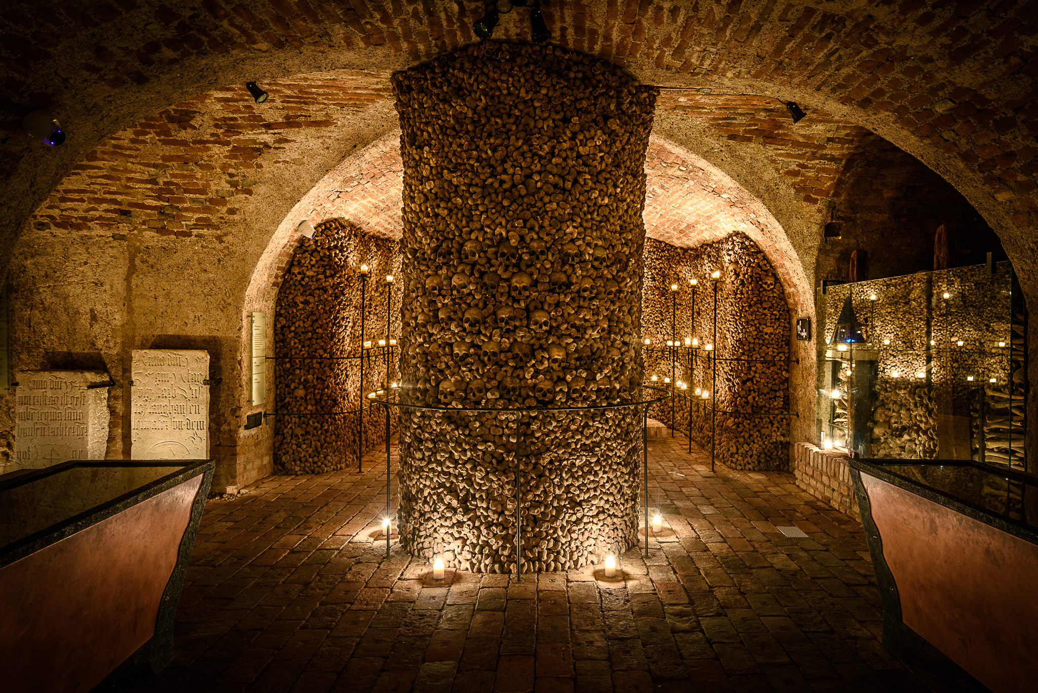 Underground ossuary with a central column of skulls and bones, surrounded by brick arches and lit by candles.