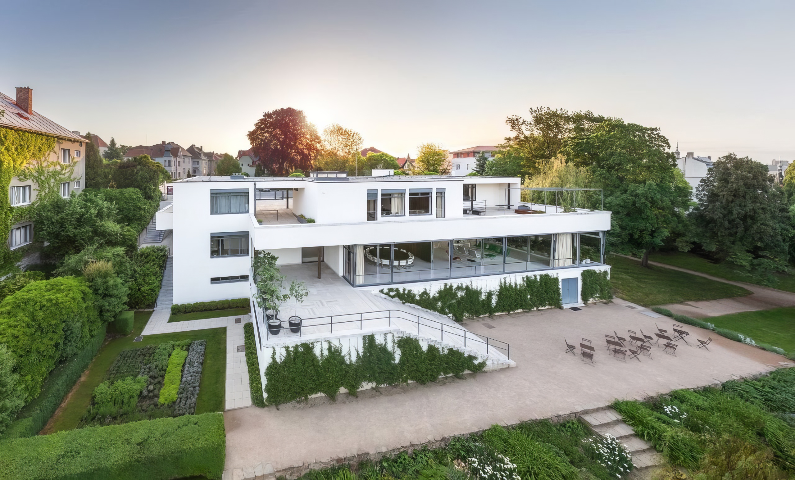 Modern white, multi-level house with glass walls and a courtyard, surrounded by lush gardens at sunset.