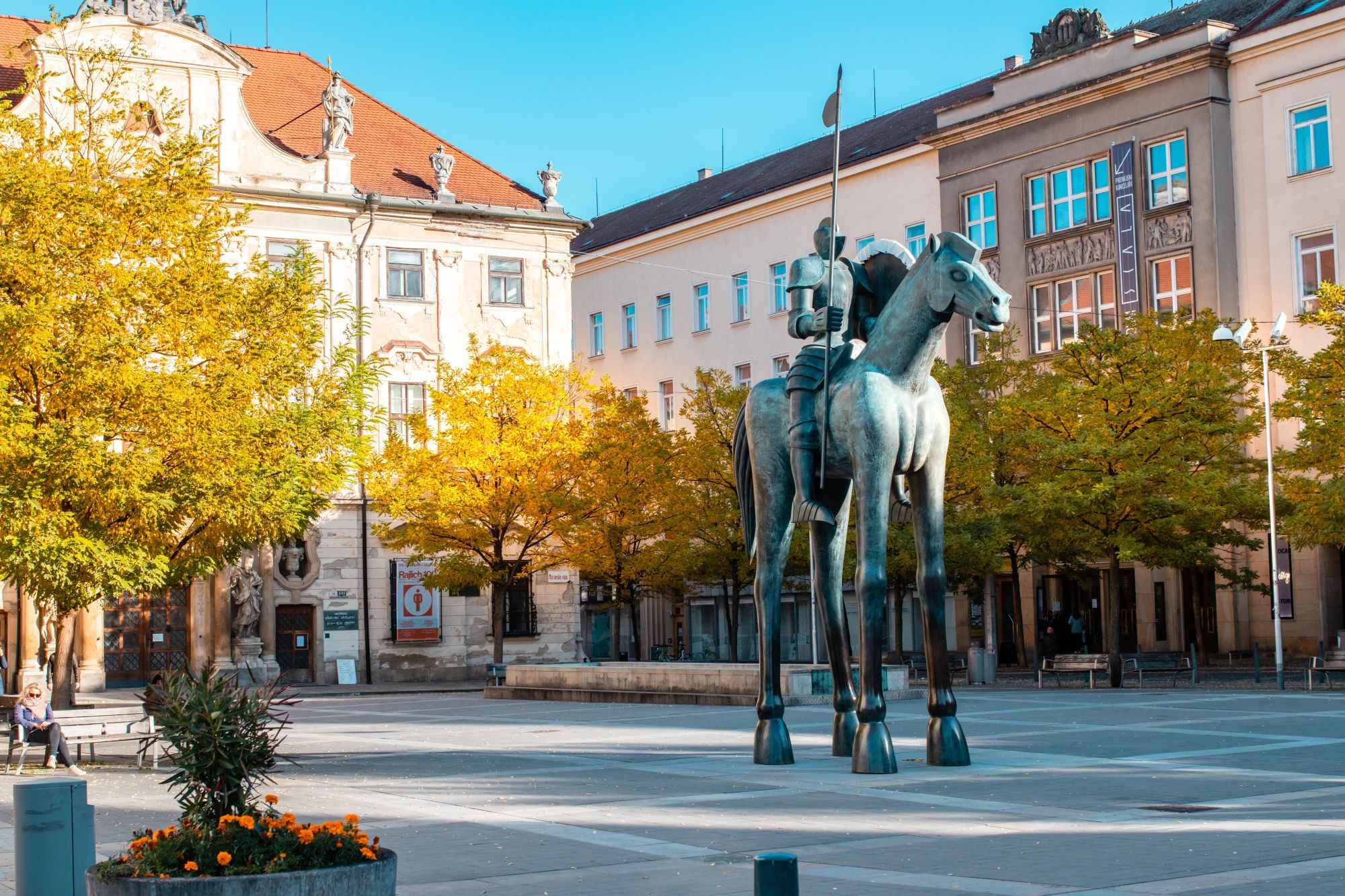 Bronze statue of a knight on a horse in a city square with autumn trees and historic buildings behind him.