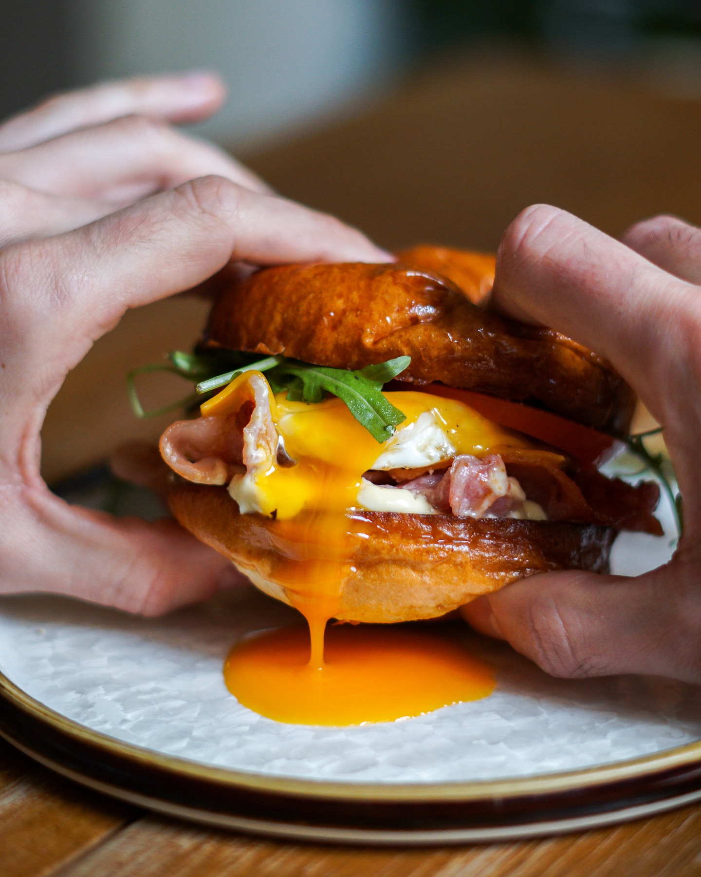 Close-up of hands squeezing a bacon cheeseburger with melted cheese, egg, arugula, and orange sauce dripping onto a plate.