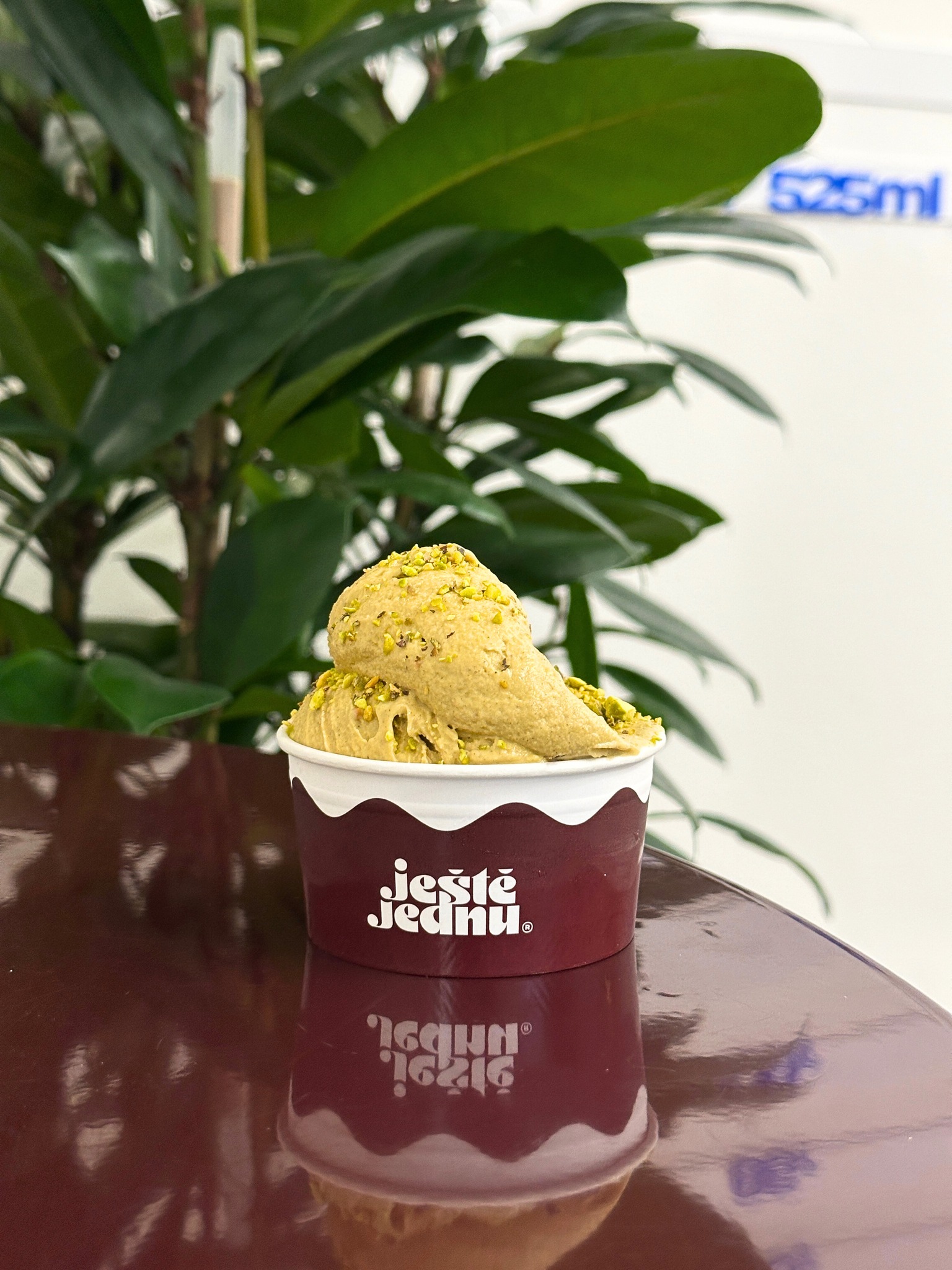 Scooped ice cream in a maroon cup with pistachio sprinkles on a table, plant in the background and white branding on the cup visible