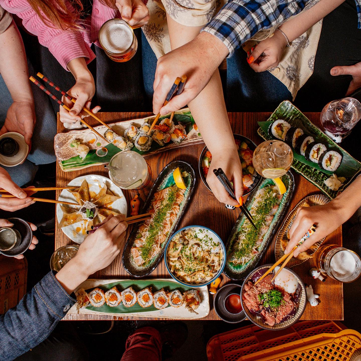 Top-down view of a shared sushi feast with several people using chopsticks to pick up pieces from assorted rolls and bowls on a wooden table.