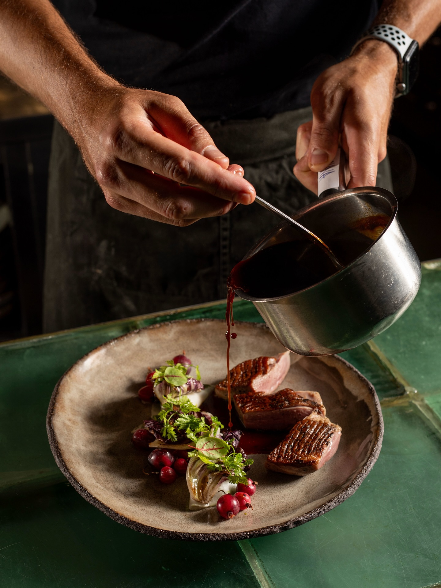 Chef pouring dark sauce over a seared steak with greens and berries on a rustic plate.