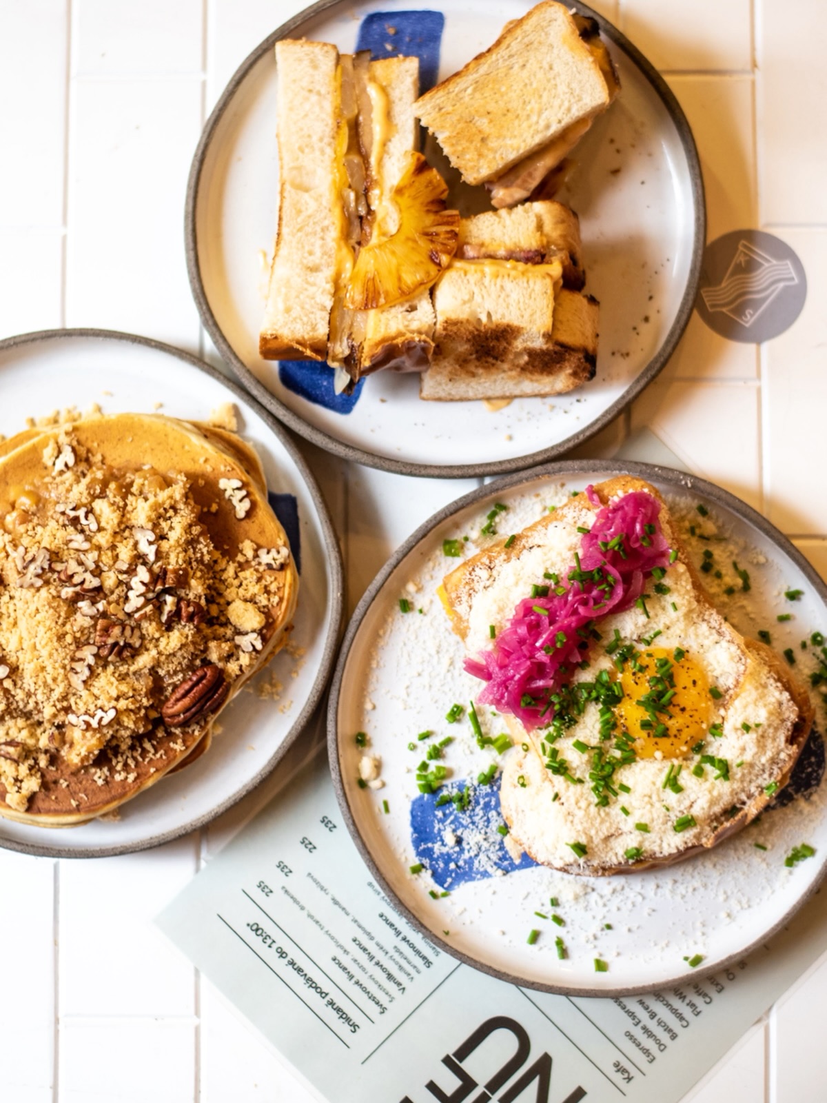 Brunch plates with an open-faced egg toast topped with pink pickled onions and chives, plus crumbly pancake and cubed grilled sandwiches on ceramic dishes.