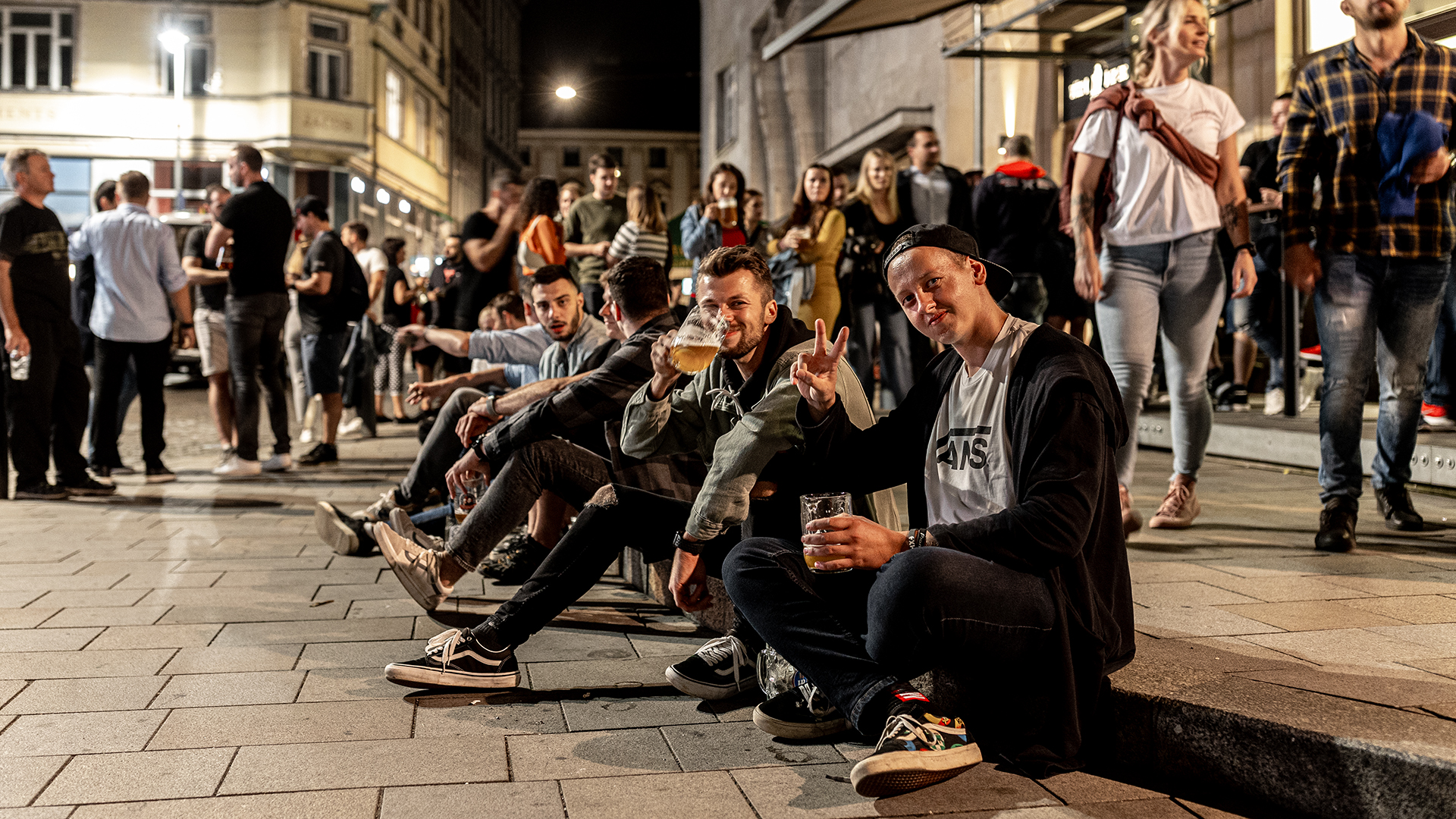 Group of friends sitting on a curb on a lively city street at night, holding beer and posing for the camera.