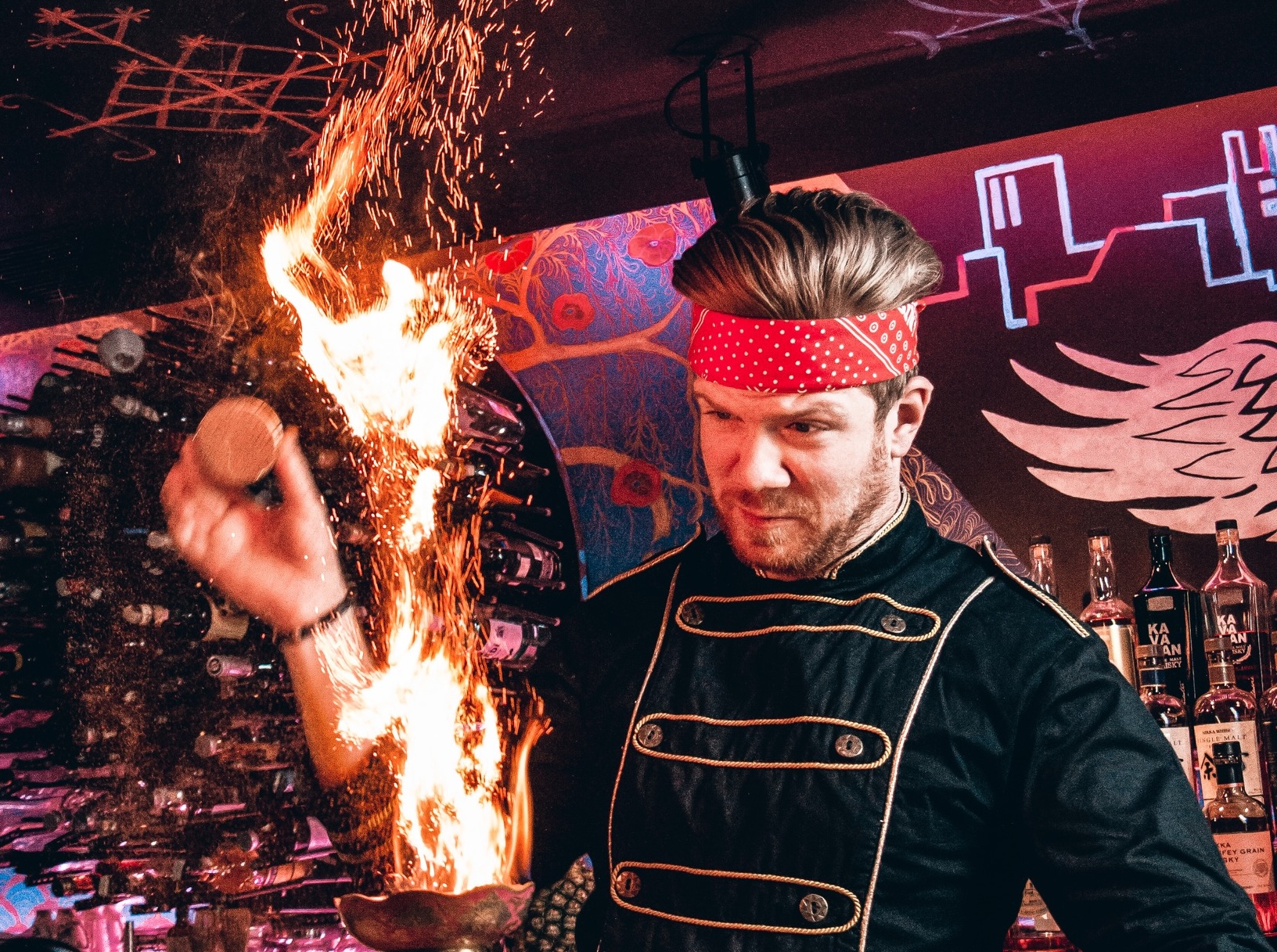 Bartender in a red polka-dot headband and navy coat performs a flaming trick, sparks flying beside a liquor bar.