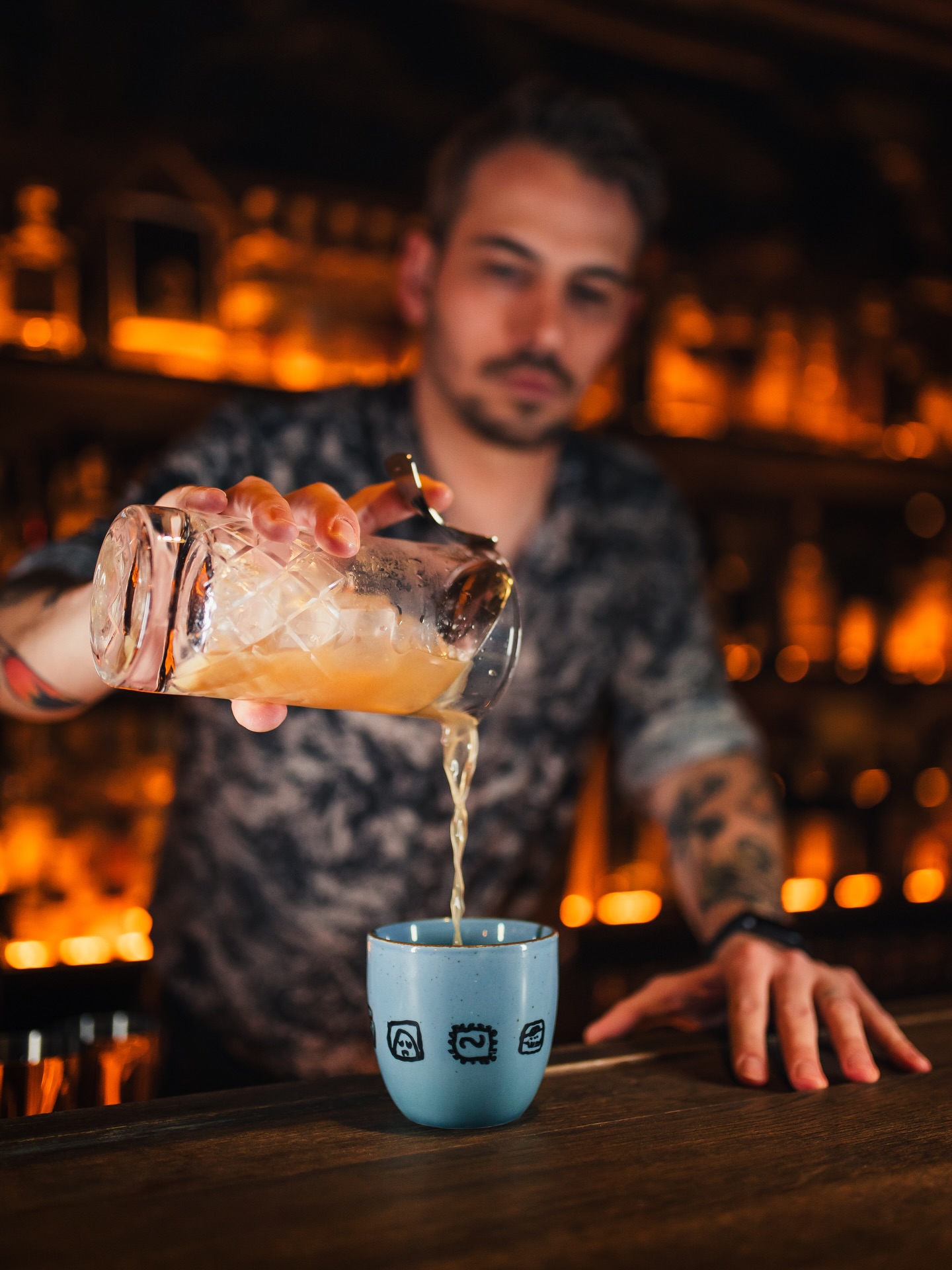 Bartender pours a cocktail from a crystal shaker into a blue mug at a dimly lit bar, ice cascading into the glass.