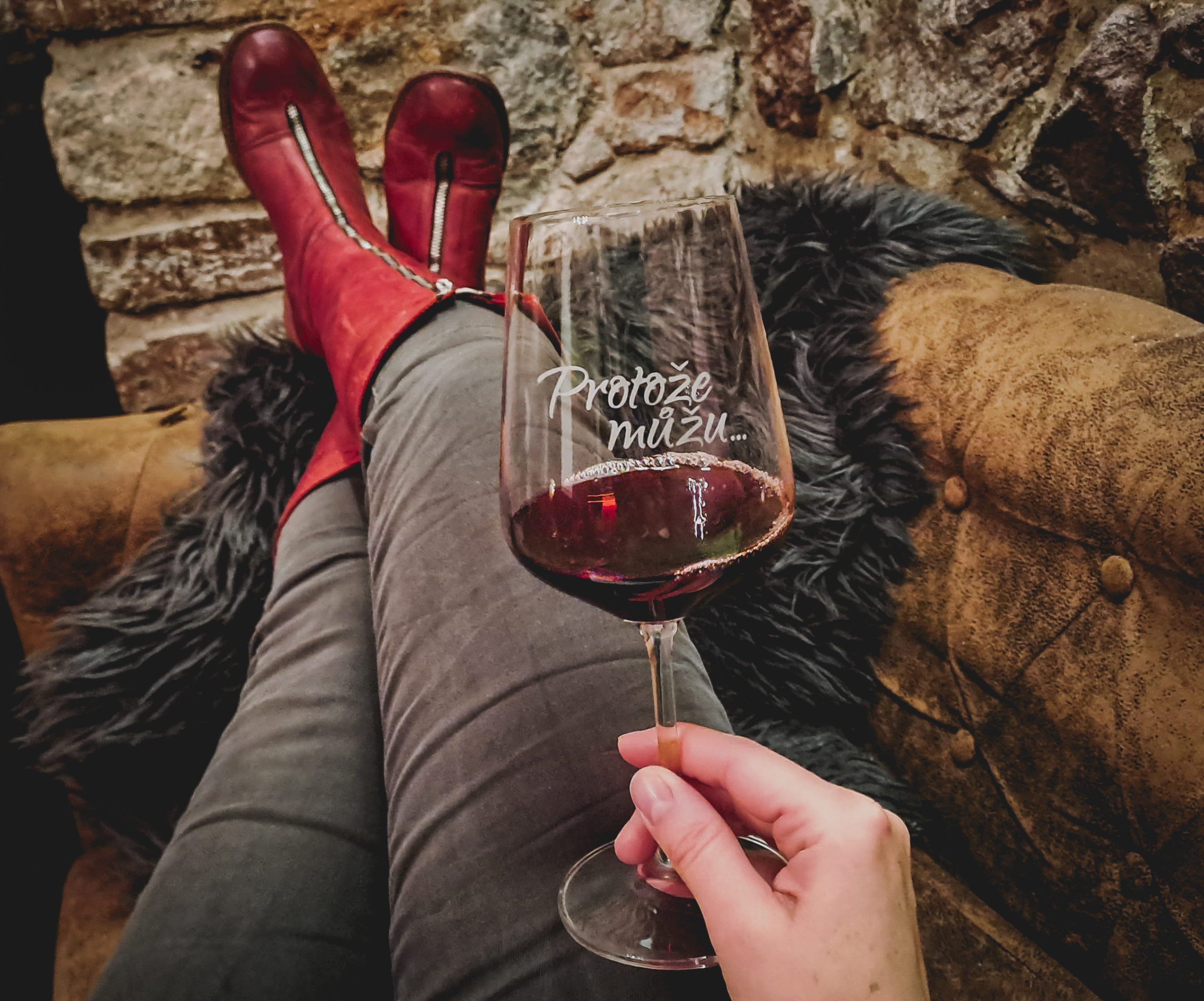 A person wearing red boots sits with legs crossed, holding a glass of red wine in a cozy rustic setting.