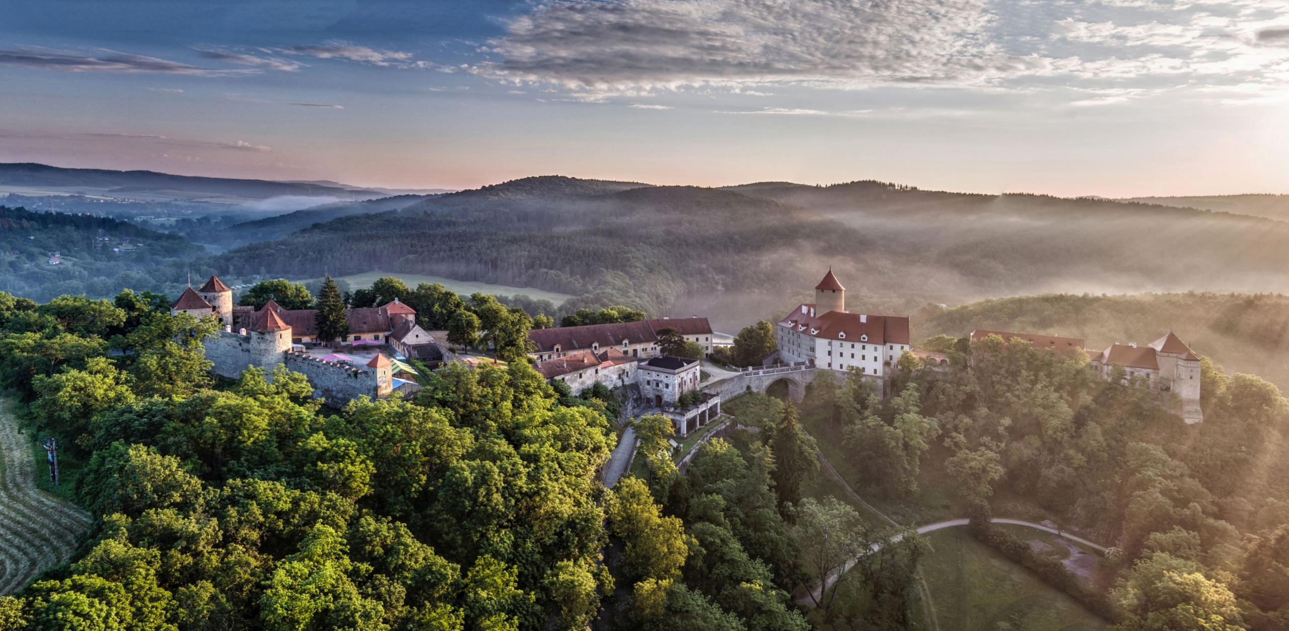 Aerial view of a medieval castle complex on a forested hill, with towers, red roofs, and a misty valley at sunrise.”] ,