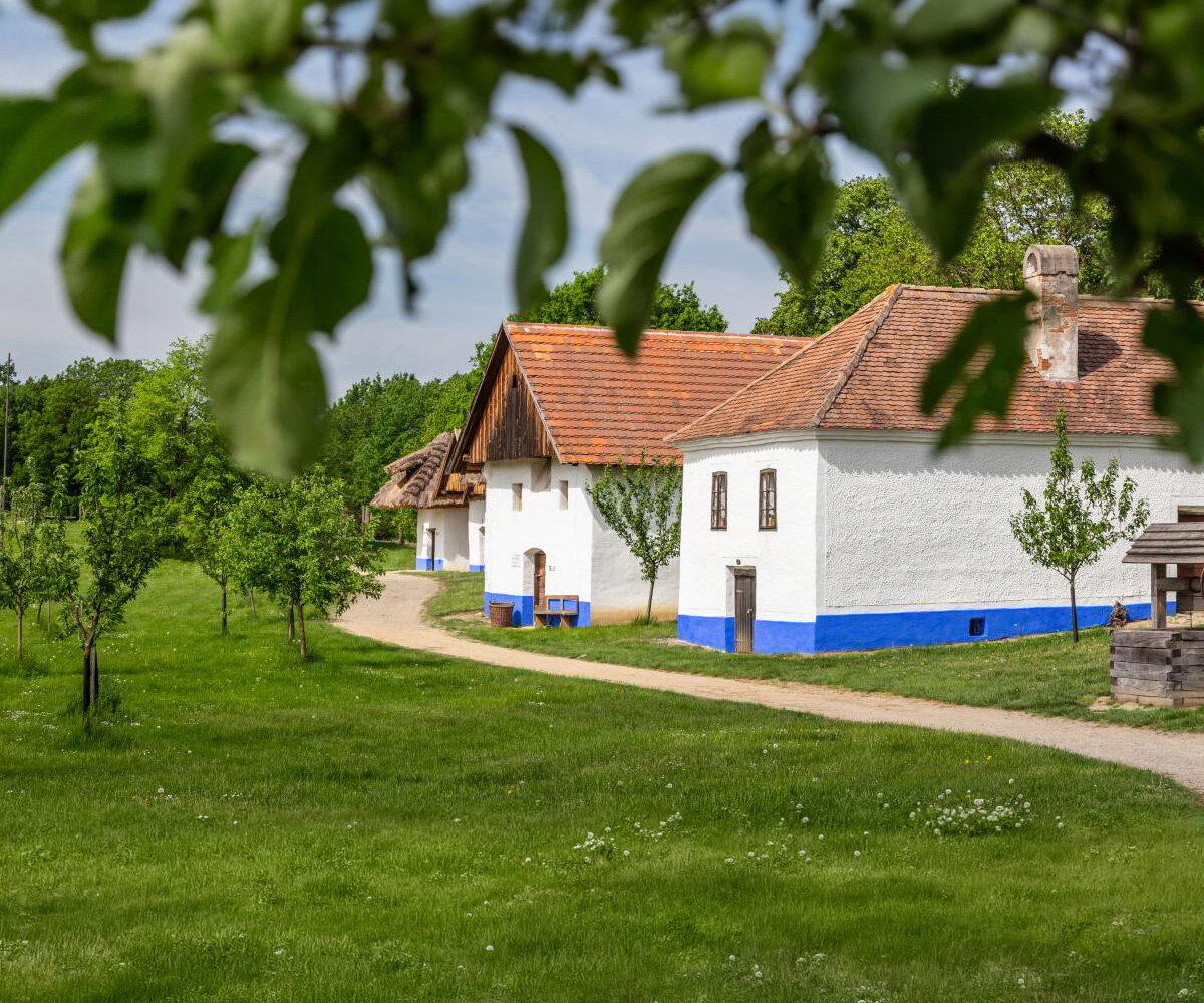 White cottages with blue bases and red-tiled roofs along a dirt path in a green park.