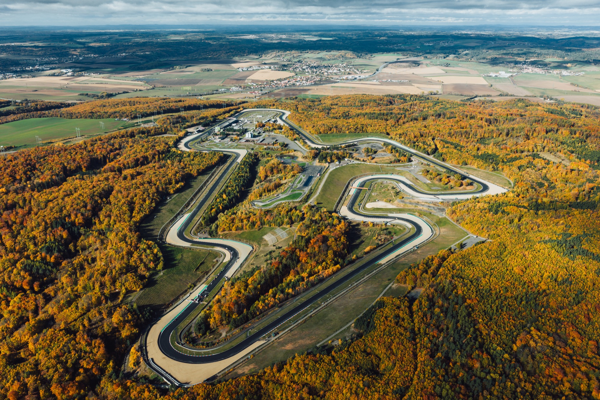 Aerial view of a winding race track through a forest in autumn colors and farmland beyond the trees in fall foliage.