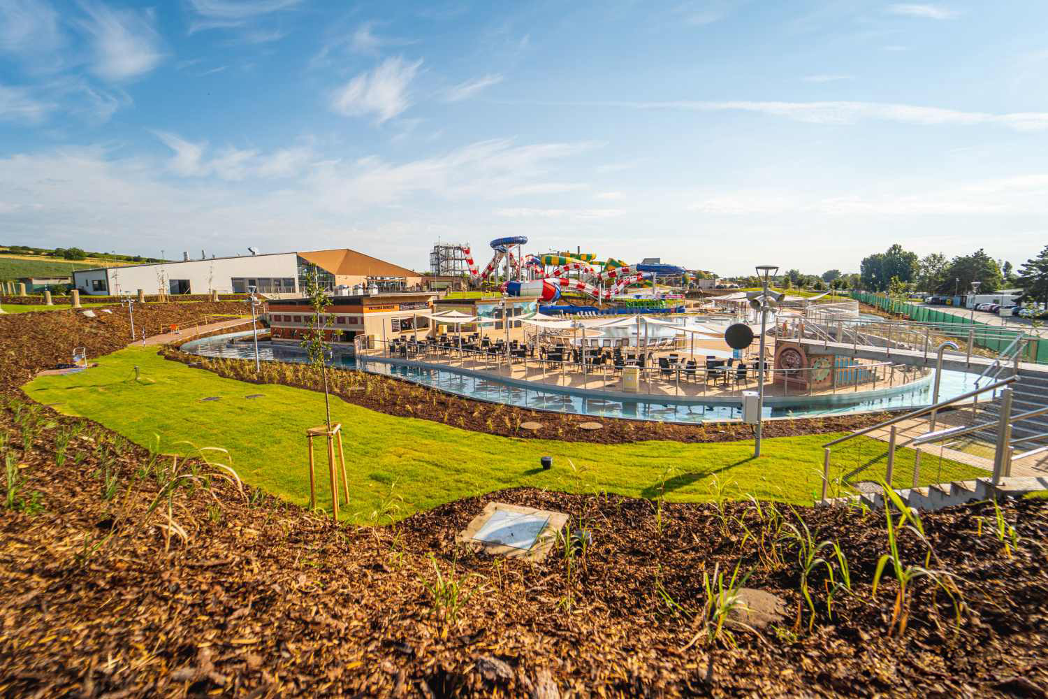 Outdoor water park with colorful slides, a circular lazy river, and seating under a clear blue sky in a landscaped area.