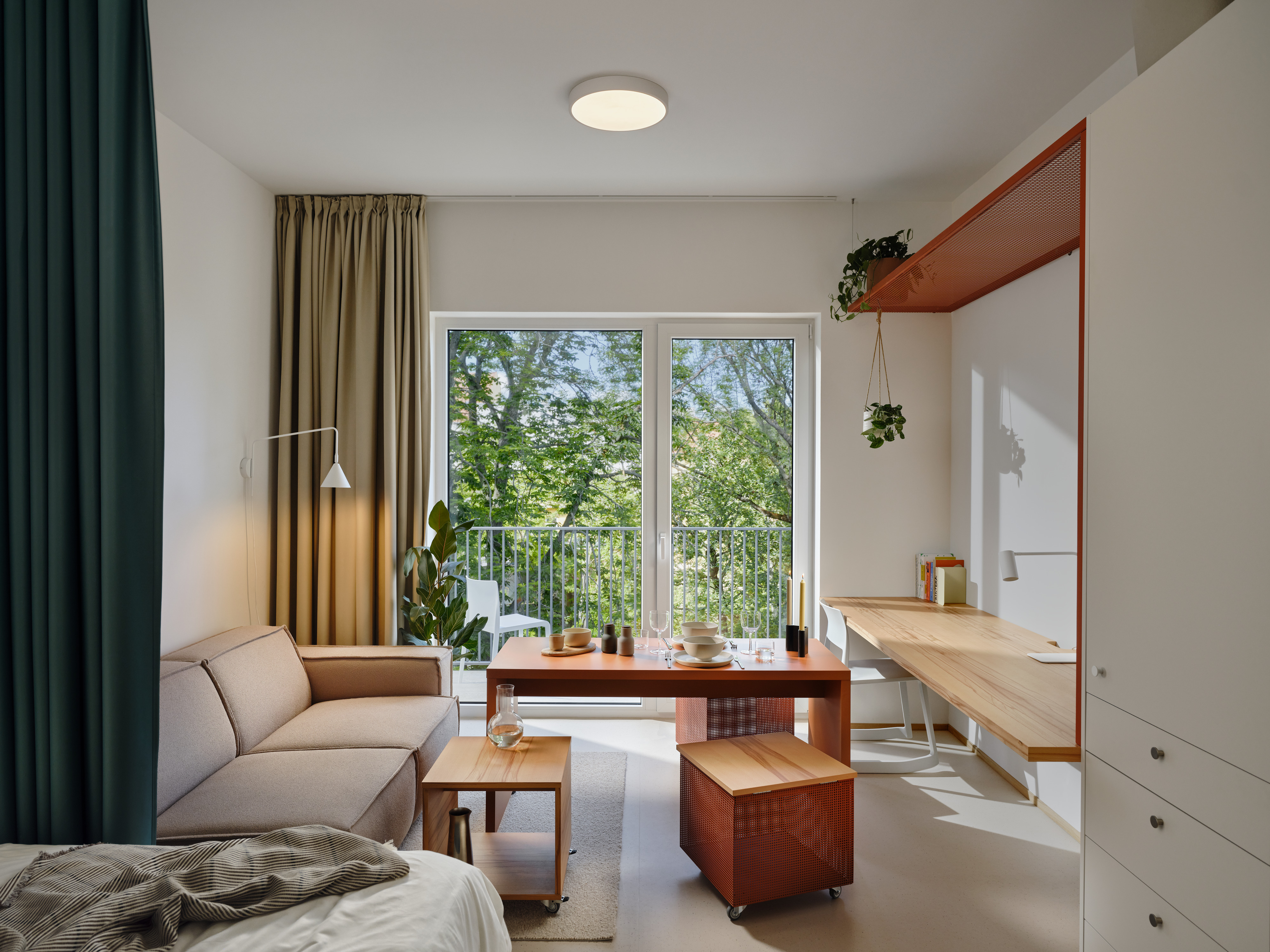 Bright living area with a beige sofa, wooden coffee table, and a dining table set near a large window showing trees outside.
