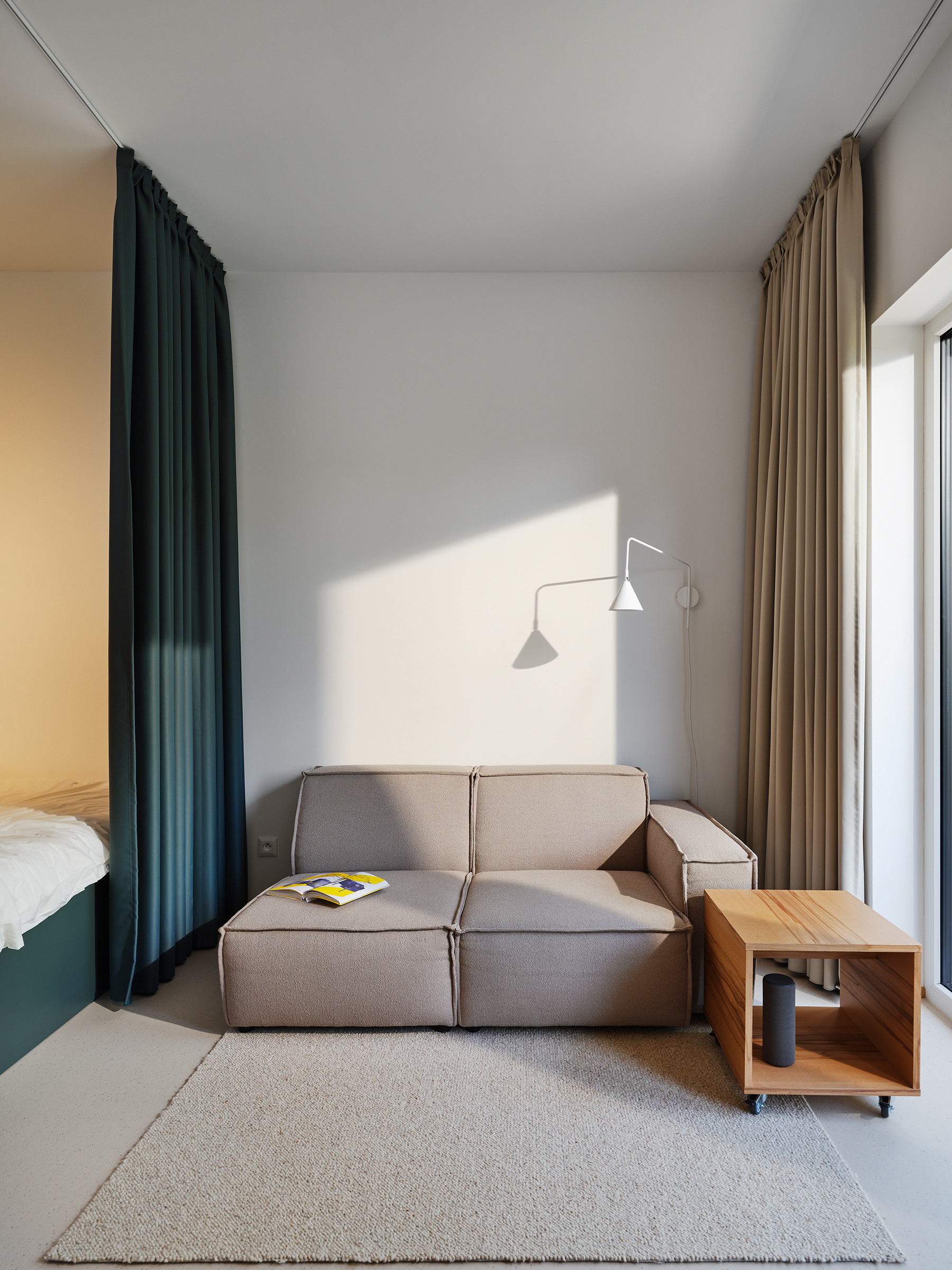 Modern living room corner with a beige sectional sofa, beige rug, and a wooden side table on wheels; dark teal and beige curtains frame a large window; wall-mounted lamp casts angled light.