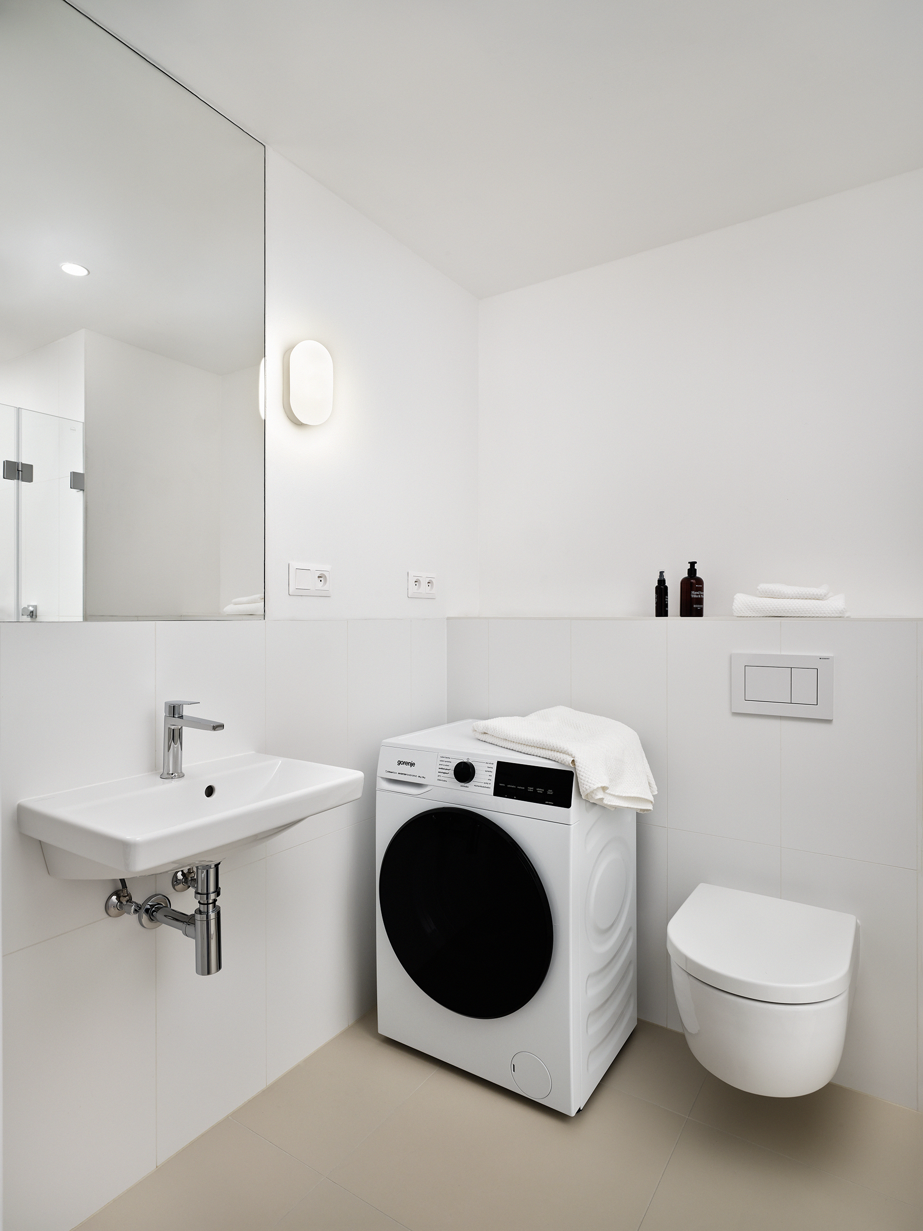 White minimalist bathroom featuring a front-loading washing machine beside a wall-mounted sink and a wall-hung toilet, with a large mirror and neatly folded towels on a ledge.
